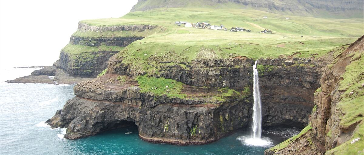The waterfall at Gásadalur falling from green cliffs into the sea, with the village on the slope above